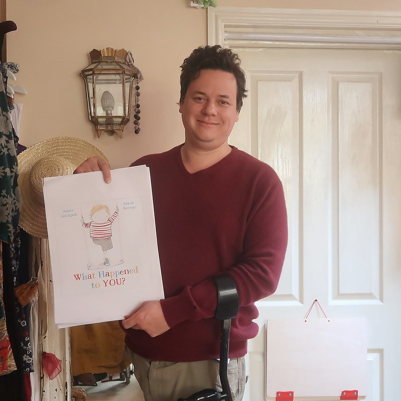 Image description: James is standing holding an early printed out version of What Happened to You? He's a one-legged white man in his late 30s, a black crutch is hooked over his arm.