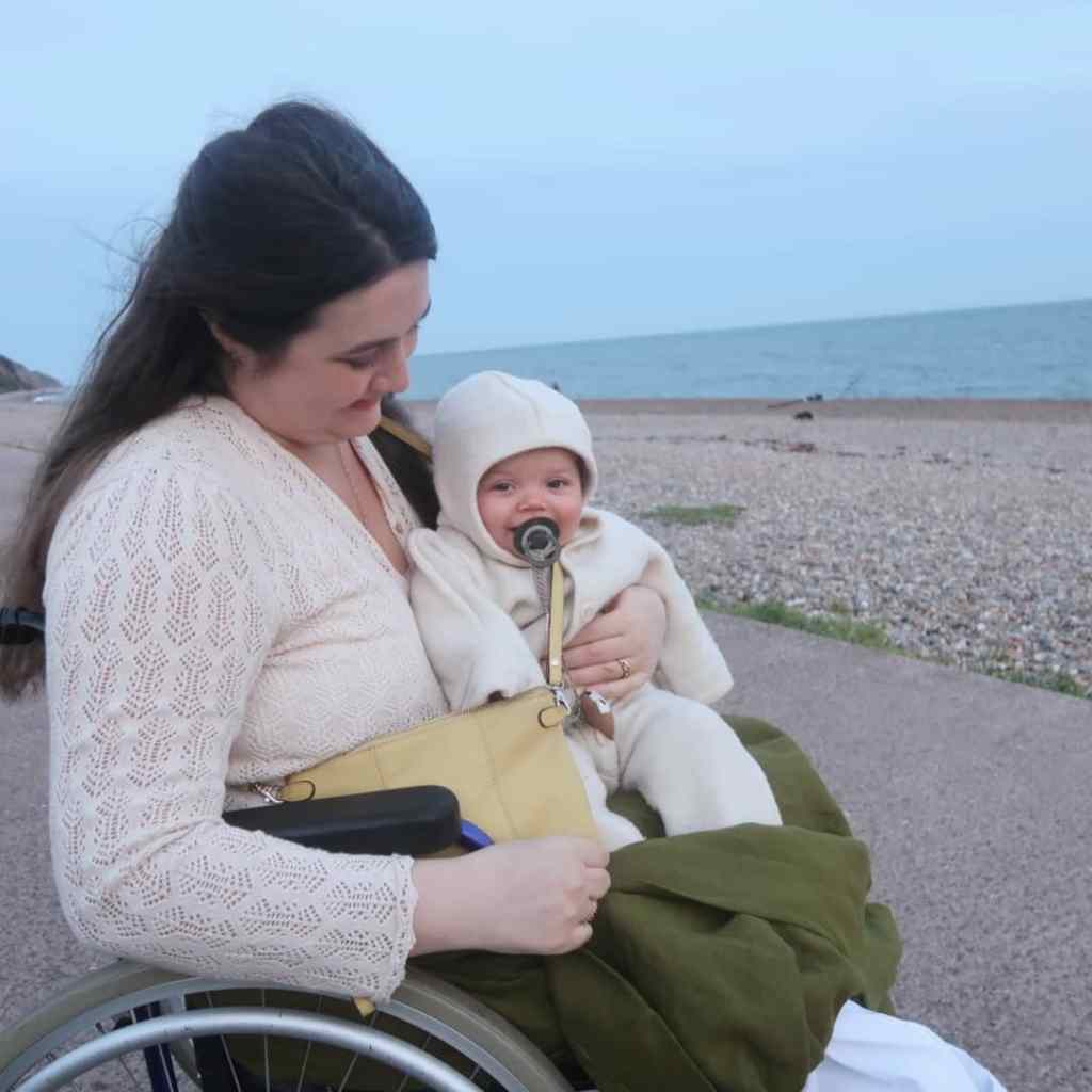 Lucy - a white woman with long brown hair - is sitting in her wheelchair on a beach. Windswept, would be a word. Her young baby Viola is sitting on her lap in a wool all-in-one, smiling at the camera. Lucy's green linen skirt is pulled up around Viola's feet to keep them warm.