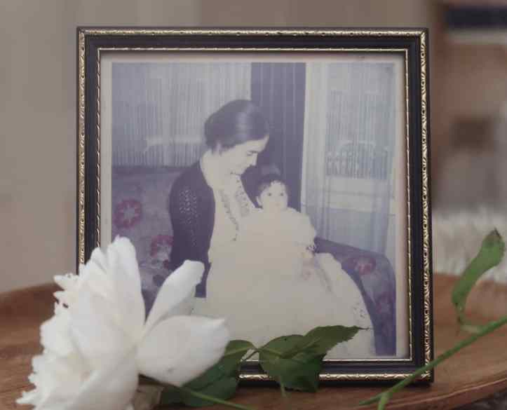 A framed photo of a young woman with dark hair and pale skin holding a very alert baby in a traditional christening dress. It does look a bit Edwardian. There's a rose lying in front of the photo.