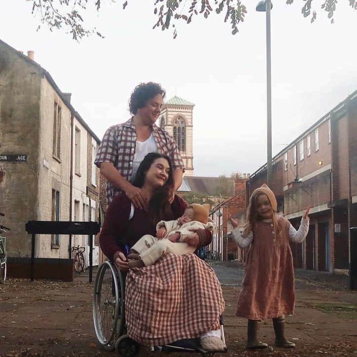 A family photo from 2019 - we're all out near our home in Oxford. Baby Viola is asleep in Lucy's lap, and Lucy is sitting in her wheelchair. Mainie (age 5) is standing to the side, gesticulating (shrugging?) and smiling at the camera. I've just realised you can't actually see that James has one leg.  (You think an entire missing limb would show up more frequently on photos to be quite honest.) Viola and Mainie are wearing matching bonnets - which are a triumph IMO. We're all white with varying shades of brown hair.