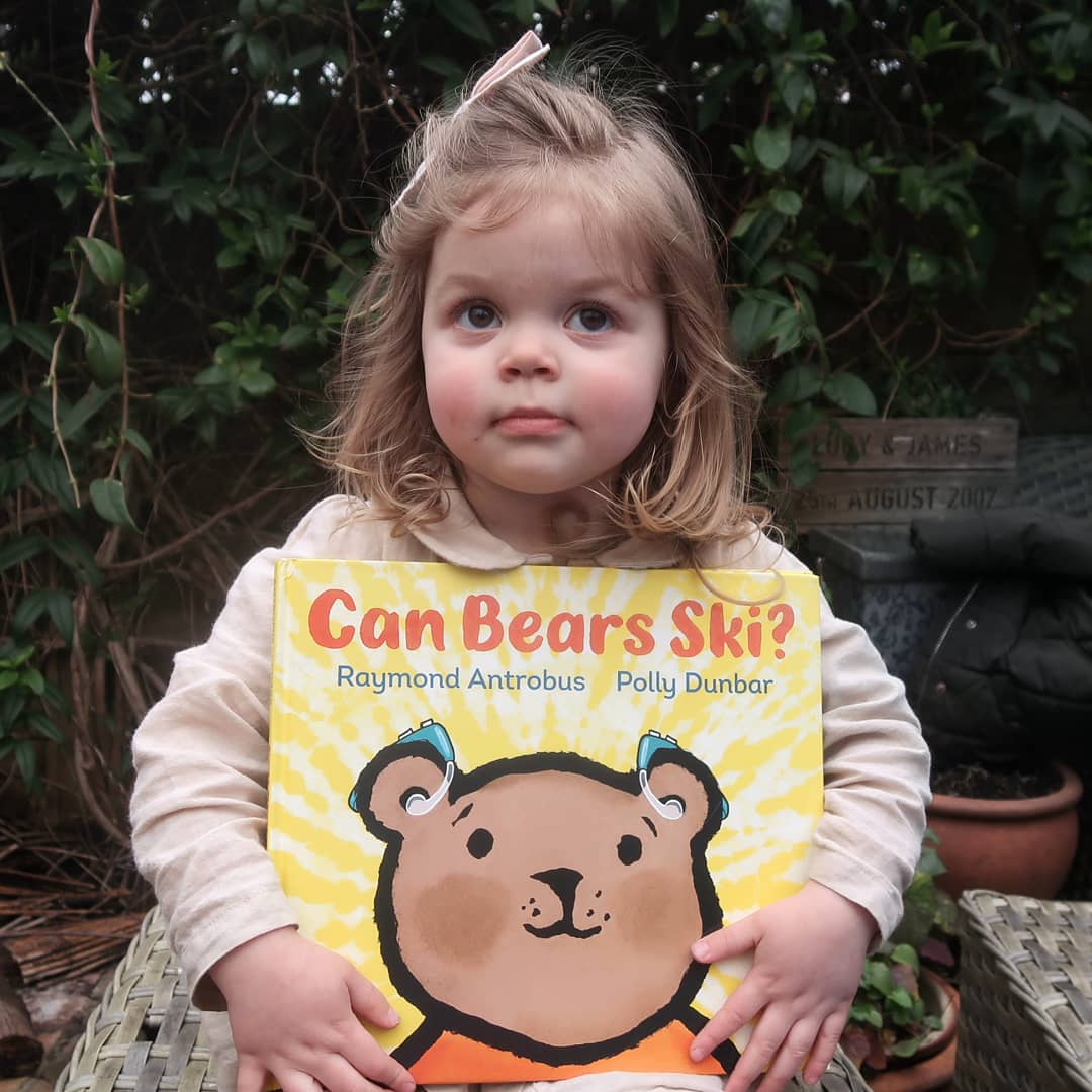 Viola sits in the garden holding a copy of Can Bears Ski? She's a 2 year old white child with light brown hair, she's sitting in front of a plant stand in our garden and looks contemplative.