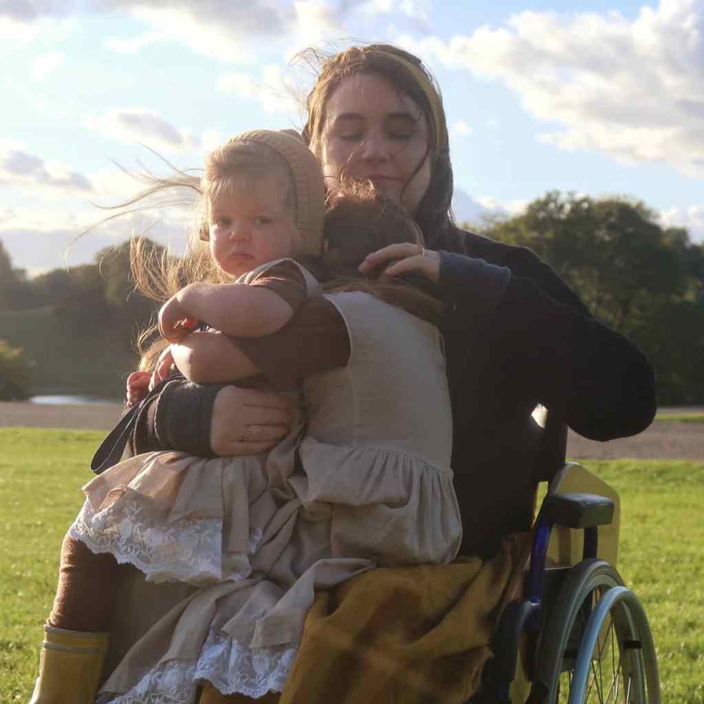 I've got both girls bundled onto my lap, you can see the top of the wheelchair wheel, and the back of a Mainie with her arms round Viola. Viola's wearing a gorgeous merino bonnet and is looking reproachfully at the camera. It's an action shot, I'm looking down and adjusting something. We're all white with light brown hair, which is blowing everywhere. I'm wearing a mustard coloured linen skirt and matching headscarf. We're in the grounds of Blenheim Palace.
