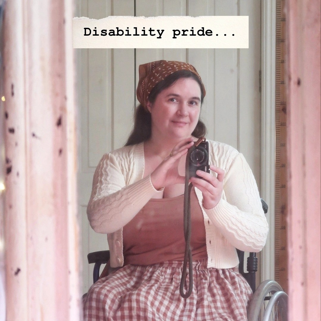 A mirror selfie. I'm a white woman wearing a brown cotton hair scarf, holding a camera and looking into the lens smiling. The words "disability pride..." are written at the top with a cream torn paper background. The mirror frame is very old and cracked, and is cream, though it looks mysteriously pinkish here.