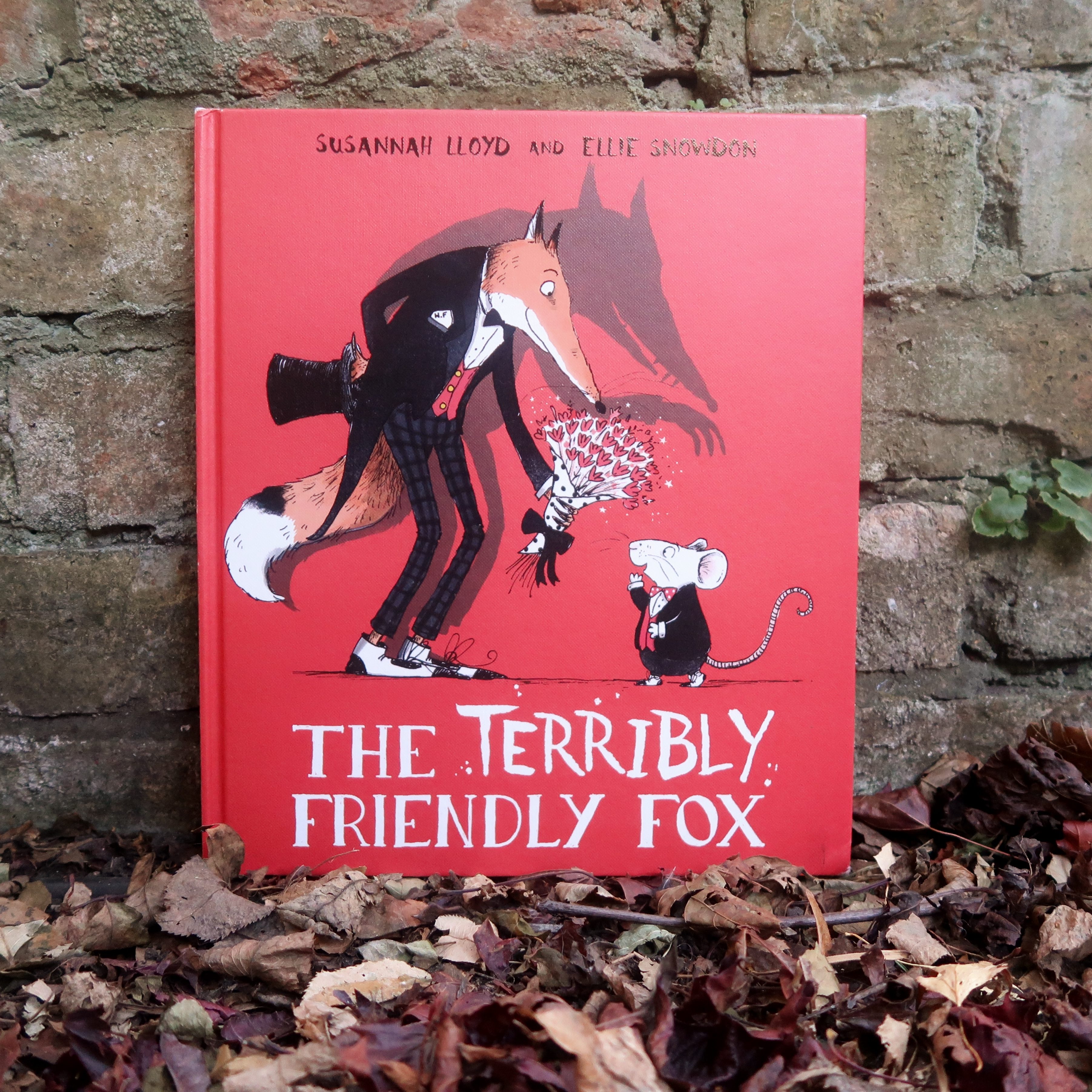The cover of The Terribly Friendly Fox - a picture book by Susannah Lloyd, illustrated by Ellie Snowden. It's a red hardback, with a besuited fox on the front, presenting a punch of flowers to a small mouse. A shadow looms behind, ominously.
