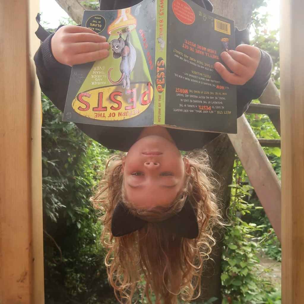 Mainie, a 6yo girl dressed as Batz, a bat, is hanging upside-down reading a copy of Return of the Pests, a middle grade novel. She's white with blondish hair and bat ears (they might be cat ears? Let's go with bat) on a hairband, and is hanging in her treehouse. It looks a bit like her eyes are closed but she is actually reading.