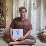 A portrait of James, a one legged white man with brown hair, holding his book - What Happened to You? He's smiling at the camera and is wearing a brown linen shirt.