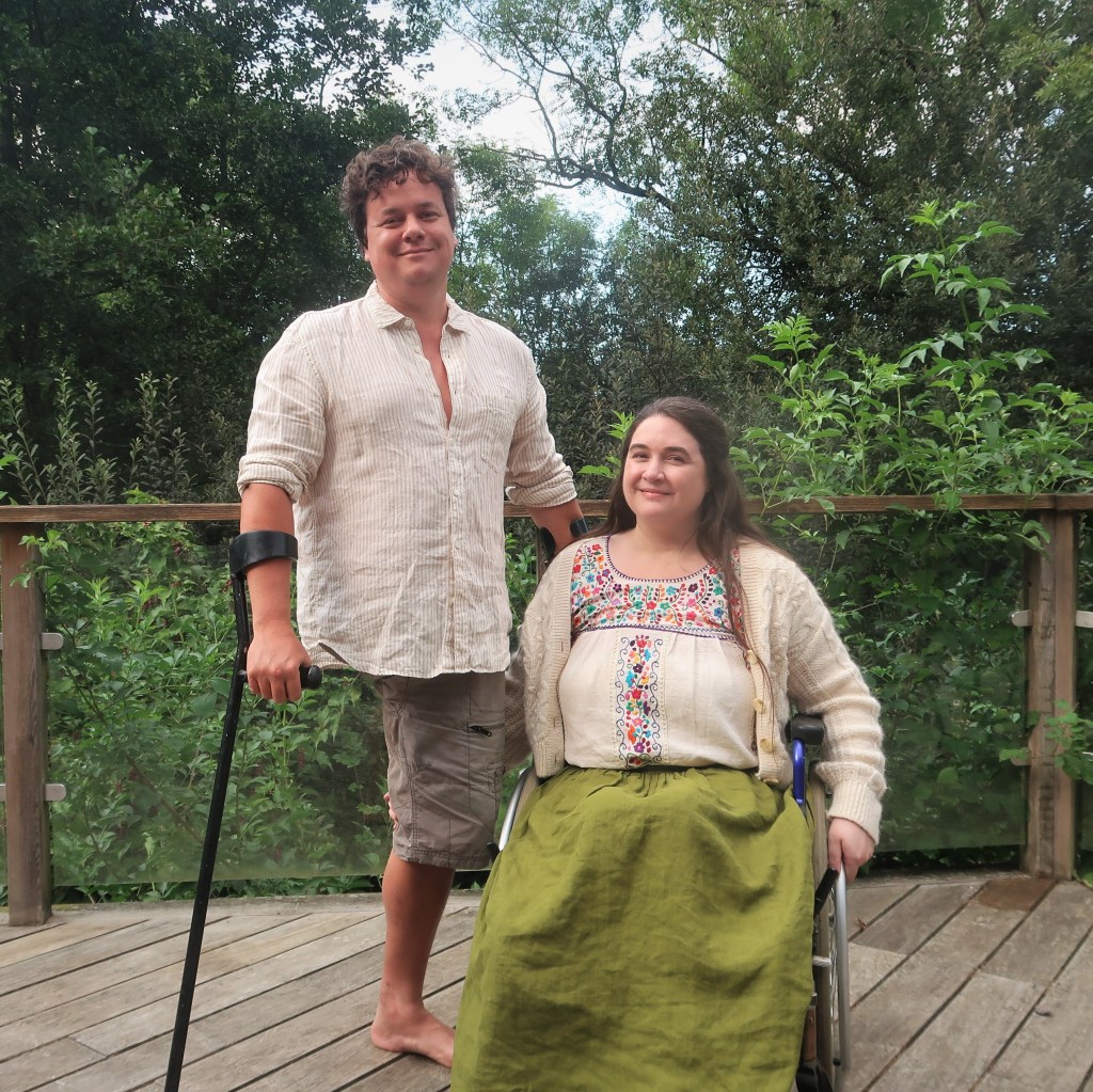 James & Lucy Catchpole. We're a white couple with brown hair. Lucy's sitting in her wheelchair, James has one leg and is using crutches.