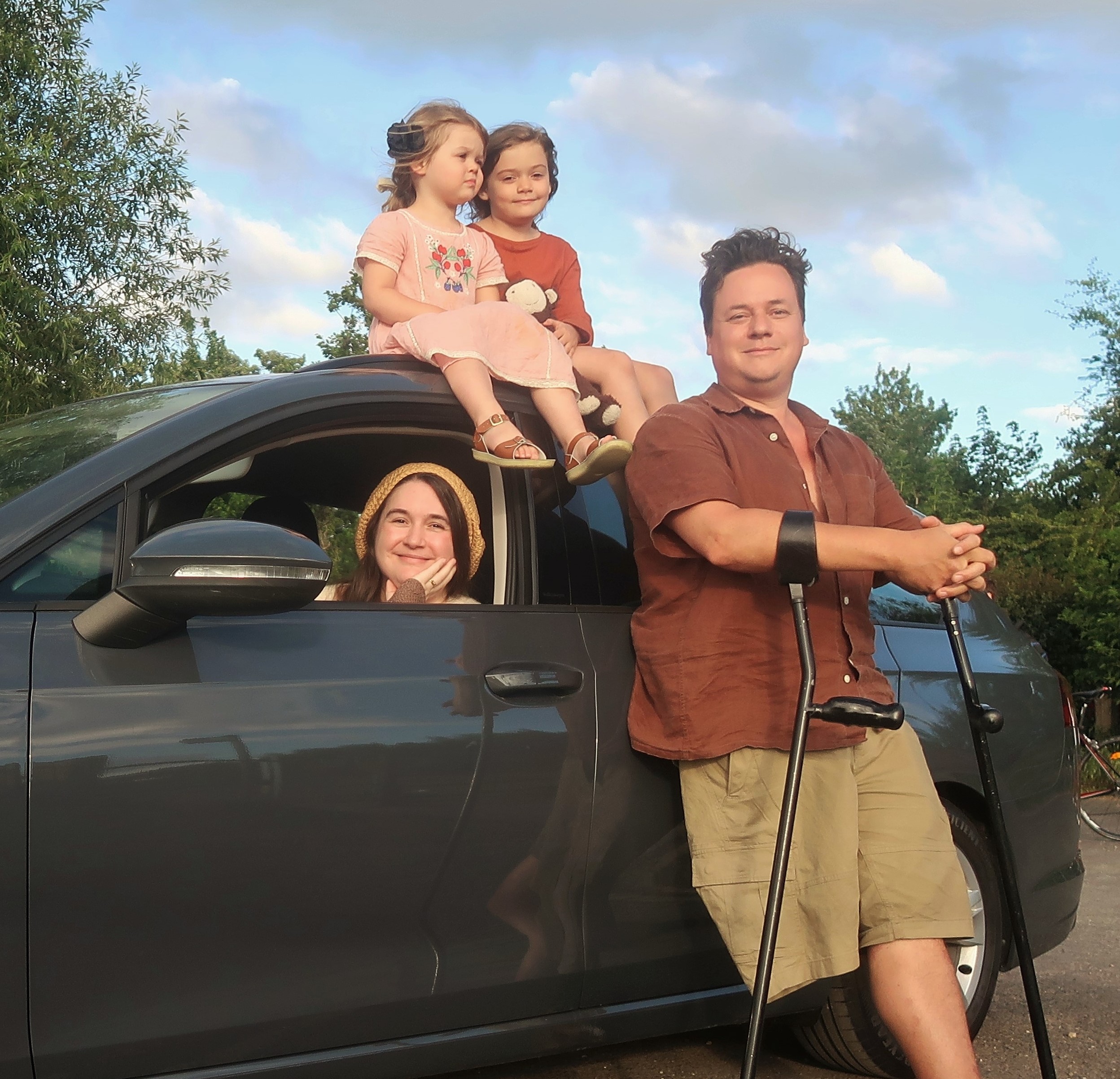 We're all with the new car. We're a white family with brown hair. Lucy's sitting in the car. James, a one-legged man on crutches, is standing in front of it. Our daughters, age 3 and 7, are sitting on the roof.