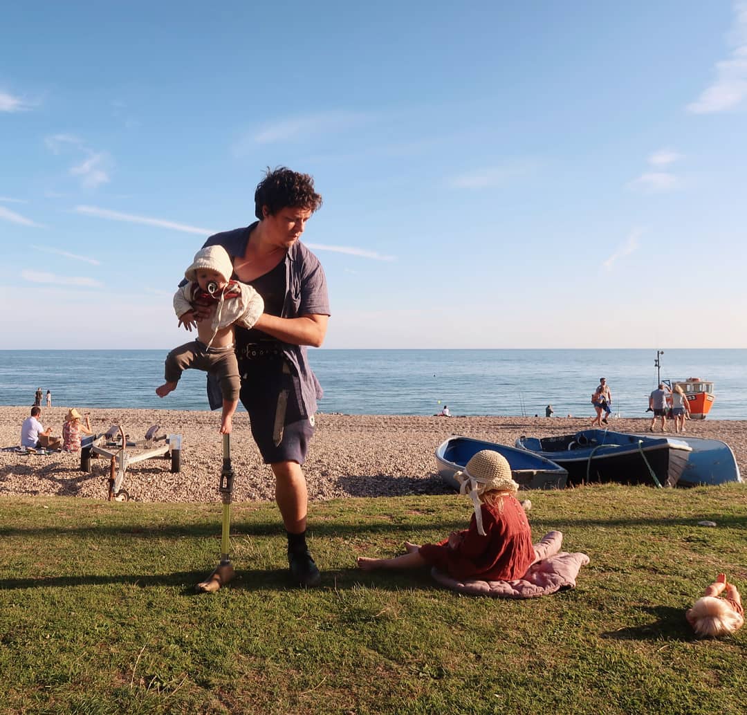 James Catchpole with young daughters. He's mid-parenting on the beach, wearing his prosthetic leg and holding his baby. His other daughter sits beside him.