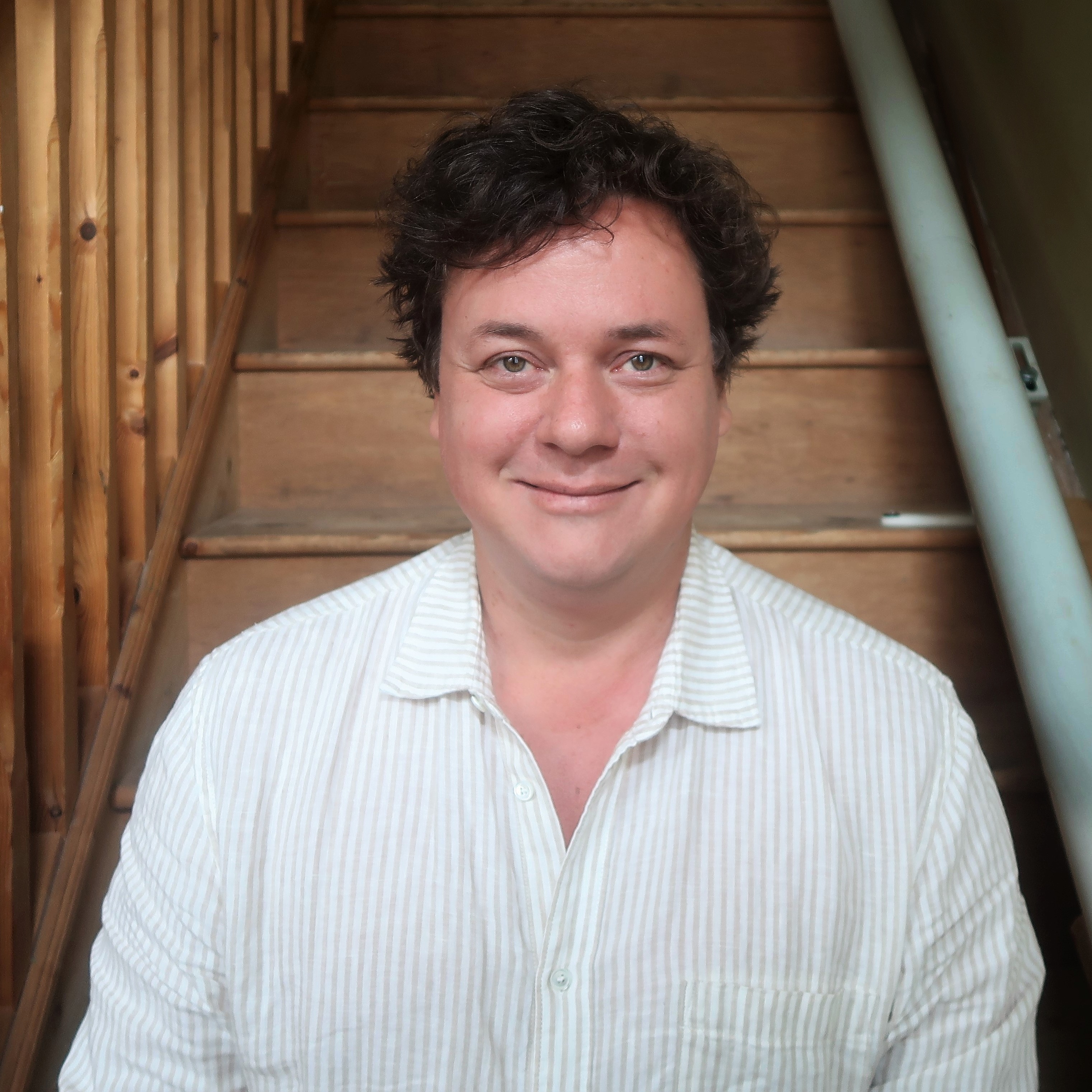 James Catchpole - headshot. A white man with brown hair, wearing a pale linen shirt. He's smiling at the camera and sitting on his stairs.