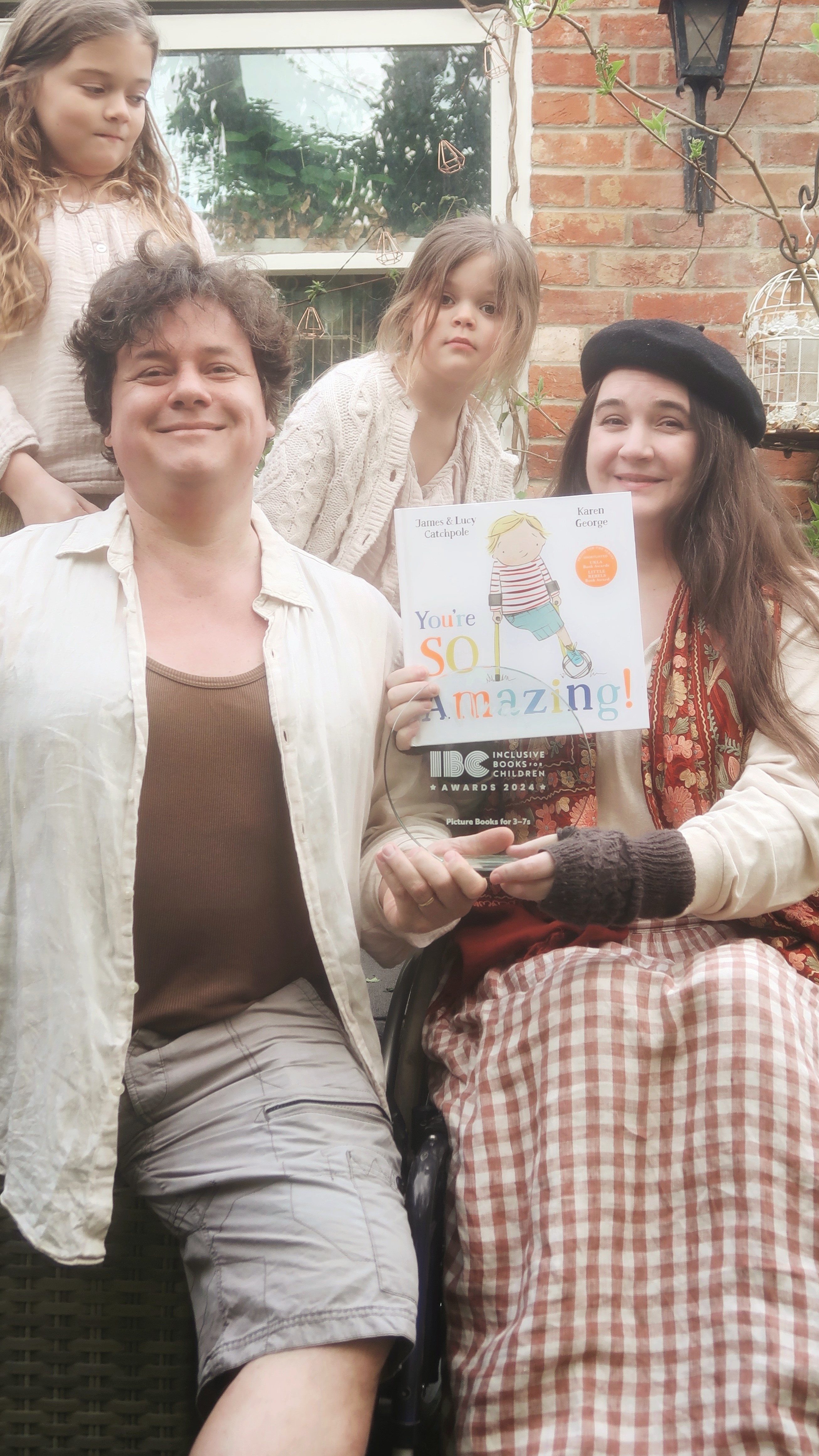 Lucy and James - a white disabled couple with their daughters - hold a copy of You're So Amazing and the IBC trophy. The trophy is transparent, making it almost invisible. Lucy is laughing.