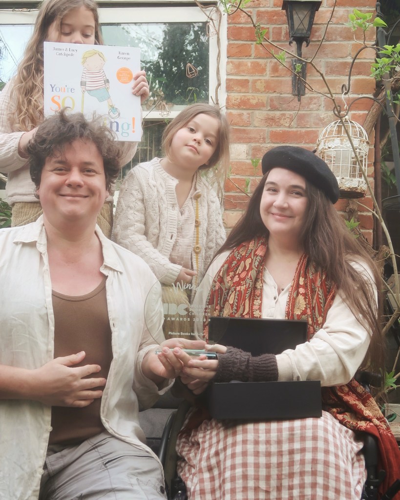 Lucy and James are sitting, holding a hard to photograph, transparent award and smiling. Mainie and Viola are standing behind on the sofa, Mainie's holding a copy of You're So Amazing!
