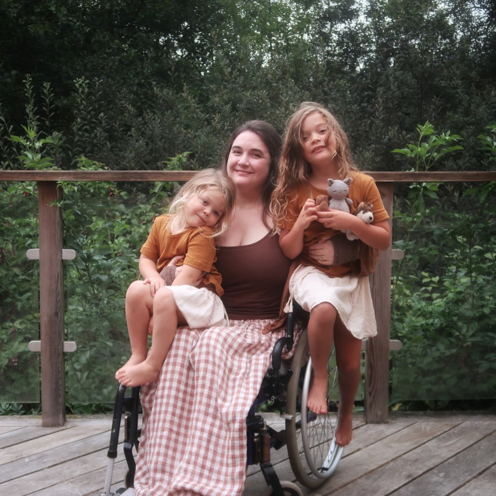 Lucy - a white woman with long brown hair - sits in her wheelchair, with both daughters perched on and around her. They're smiling at the camera.