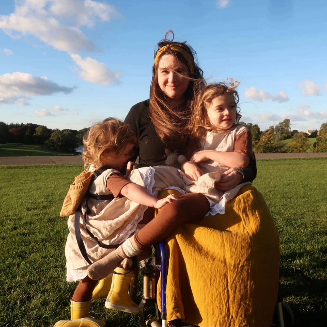 Lucy Catchpole and her daughters - a white mother with long brown hair, wearing a honey-coloured linen skirt, she's outdoors in her wheelchair. One daughter is on her lap while the other is trying to climb aboard.