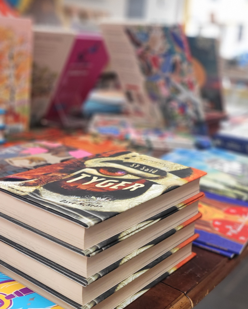 Tyger. A stack of hardbacks of Tyger by SF Said, prominently displayed on a wooden table. The cover art is a striking image of an eye and part of a face.