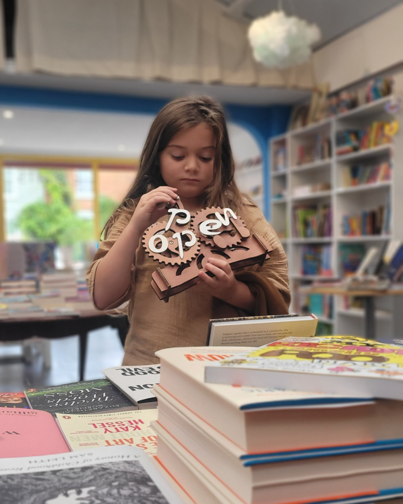 The bookshop open and closed sign Mainie’s holding a wooden mechanical puzzle shaped like gears - the word it makes changes from ‘closed’, to ‘open’.
