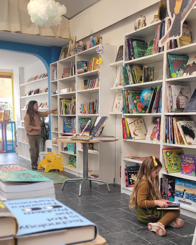 Mainie and Viola browsing the bookshop. Viola sits on the floor reading a book, Mainie is browsing the shelves. A white cloud-,like decoration hangs from the ceiling, and there’s a large window with lovely natural light.