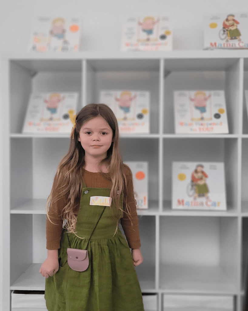 Viola, with a display of our picture books. White display shelves in the event room of Caper, with copies of You’re So Amazing! What Happened to You? and Mama Car. The first two books have a one-legged child on the cover. (Illustrated by Karen George.) Viola (5) is white with long brown hair, a yellow bow on the side. She is wearing a green linen pinafore with a brownish-orangey top underneath. She’s smiling at the camera.