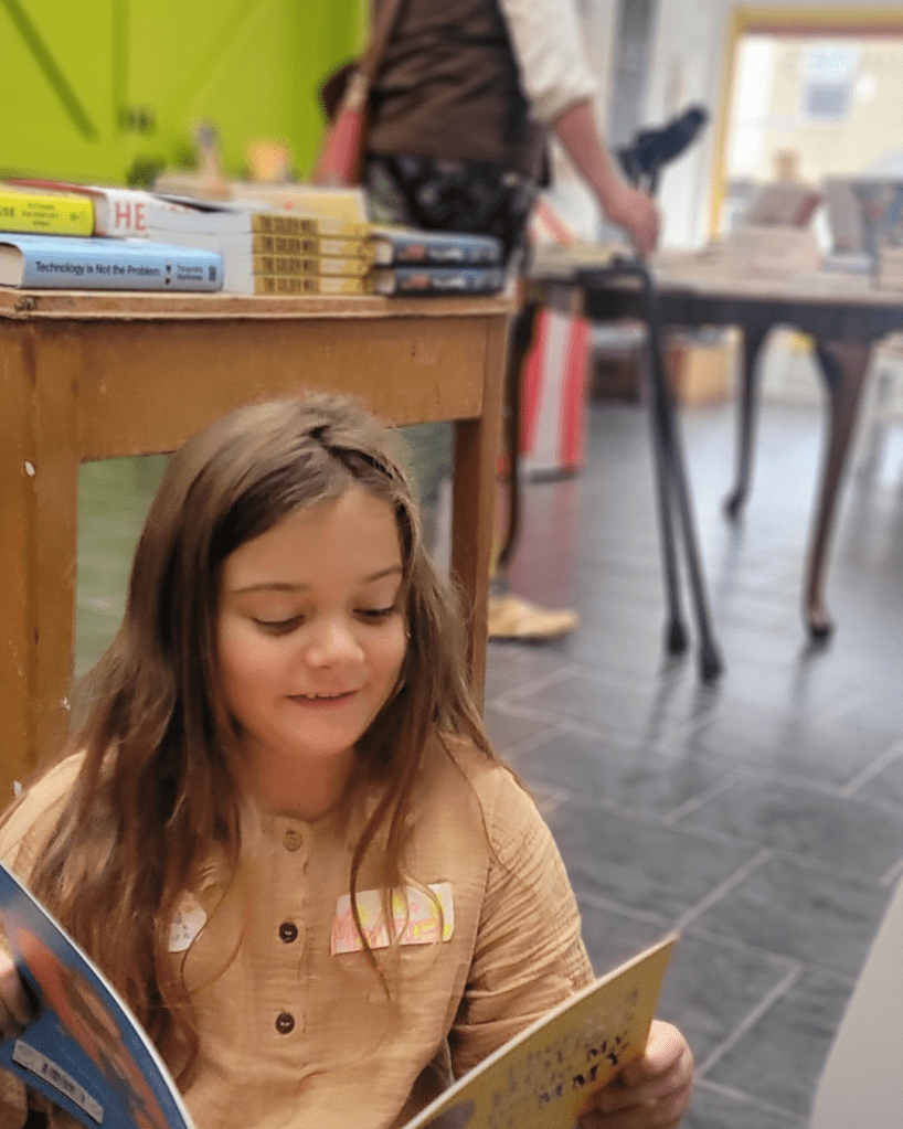 Mainie, reading in the bookshop. Mainie sits cross-legged on the floor, laughing and reading a book. She has a name tag on - which she made. James and his crutches are blurred in the background.