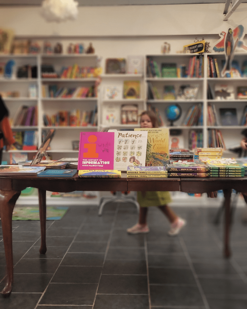 Patience Viola, out of focus, walks behind table piled high with picture books - standing up in the middle is Patience, by Rachel Williams and Leonie Lord. The table is made of dark wood with elegant legs.