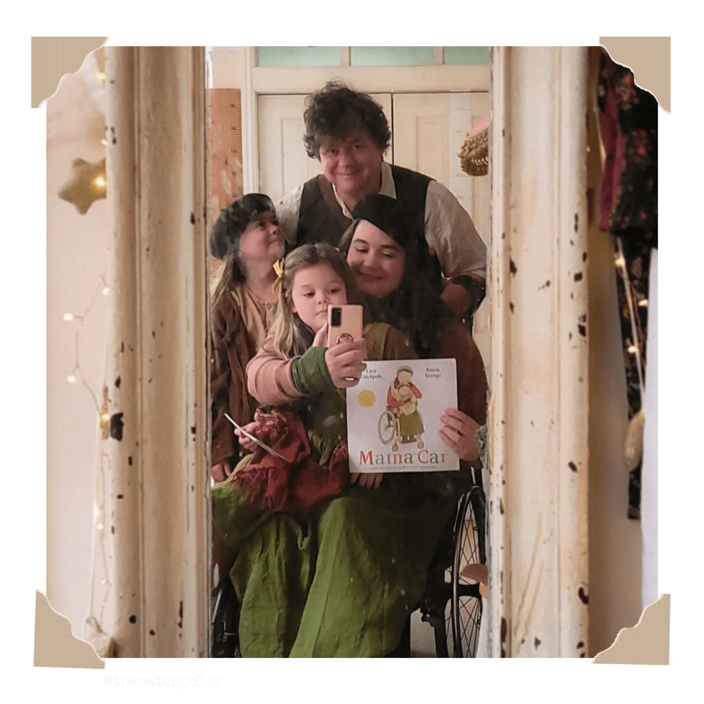 A mirror selfie of Lucy and James Catchpole, back in our house. I’m in my wheelchair, holding a book and taking a selfie with my phone. Viola’s sitting on my lap, while Mainie and James stand behind. The mirror is ancient and worn, with a white painted frame. The room behind is warm and light.