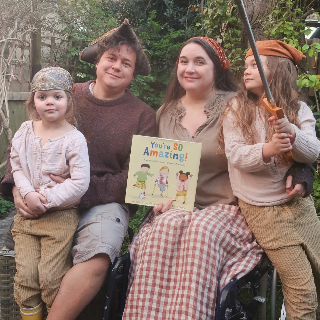 A photo of our family, celebrating "You're So Amazing!" receiving a Schneider Family Book Award Honor from the ALA. We’re in our garden, trees and greenery behind. James and Lucy - a white disabled couple - and their two young daughters wear clothes that vaguely suggest a pirate theme, with hats and bandanas. We’re all sitting, with Lucy in her wheelchair - holding our picture book "You're SO Amazing!". Lucy and James are smiling at the camera. Book cover: “You're So Amazing” - the US hardback, a children's picture book with a cream background. On the illustrated cover are three children between 4 and 6yo - an East Asian boy, a white one-legged boy with yellow crutches, and a Black girl. They have a football and look happy. 