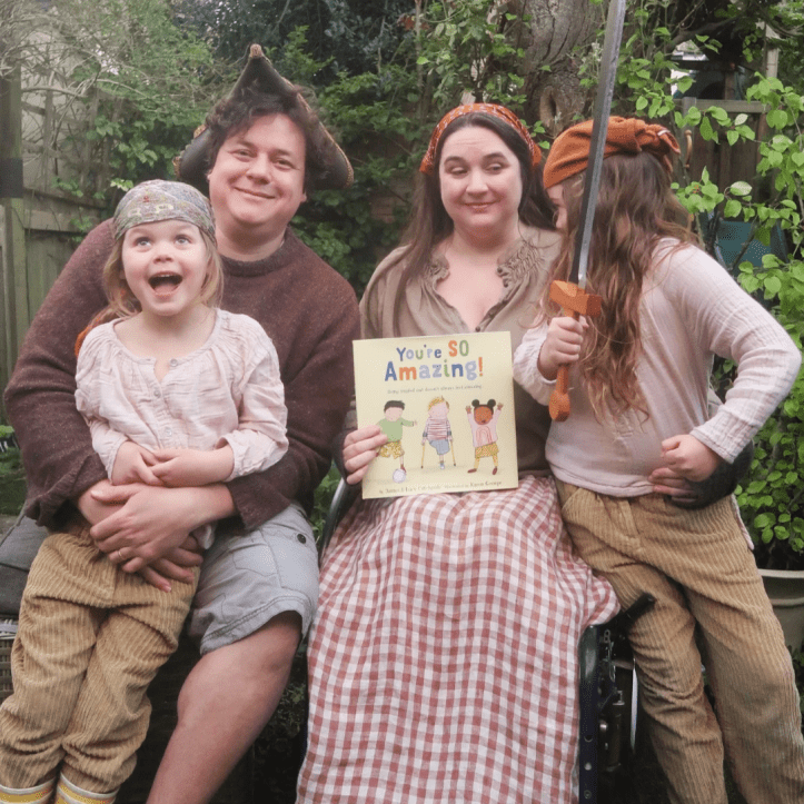 A photo of our family, celebrating "You're So Amazing!" receiving a Schneider Family Book Award Honor from the ALA. Text reads: “Our book about not being amazing has a Schneider Family Book Award Honor! And to celebrate, we have a couple of books…” We’re in our garden, trees and greenery behind. James and Lucy - a white disabled couple - and their two young daughters wear clothes that vaguely suggest a pirate theme, with hats and bandanas. We’re all sitting, with Lucy in her wheelchair - holding a book our picture book "You're SO Amazing!". Viola - age 6 - has an open-mouthed expression of great joy / surprise - who can tell? Book cover: “You're So Amazing” - the US hardback, a children's picture book with a cream background. On the illustrated cover are three children between 4 and 6yo - an East Asian boy, a white one-legged boy with yellow crutches, and a Black girl. They have a football and look happy. The title "You're SO Amazing!" is in large, bold, colorful, and stylized letters in yellow, orange, blue, and light green. The tagline is "Being singled out doesn't always feel amazing". At the bottom of the cover, the credit line "By James & Lucy Catchpole • Illustrated by Karen George" in a smaller typeface.