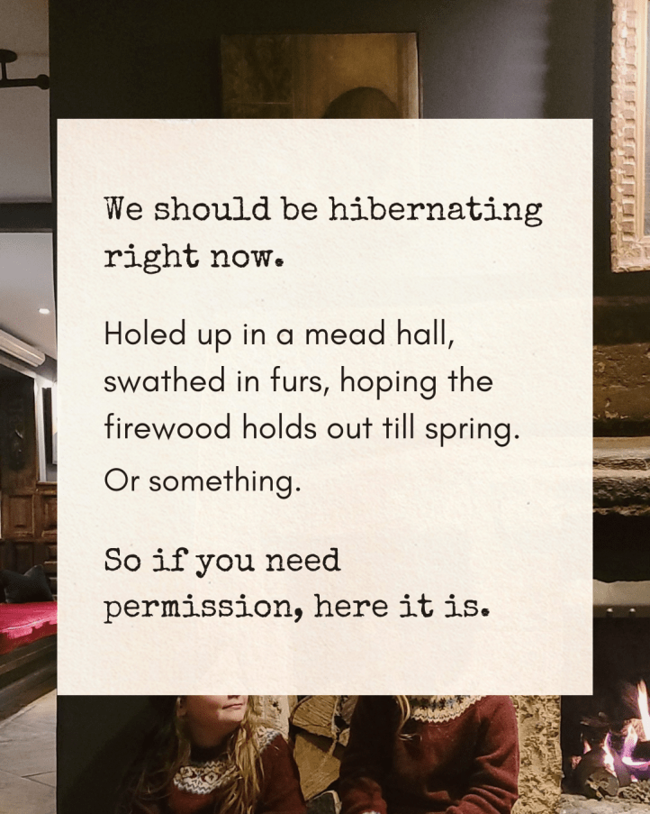“We should be hibernating right now. Holed up in a mead hall, swathed in furs, hoping the firewood holds out till spring. Or something. So if you need permission, here it is.” The background photo is slightly out of focus, you can see a dark-toned wall, a fireplace with a log fire, and a part of a red upholstered bench. A small portion of the girls in winter jumpers are visible in the background. A cosy, wintry atmosphere.