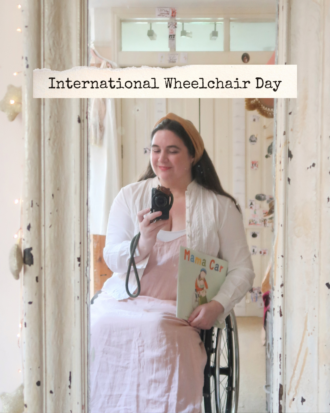 Text reads: ‘International Wheelchair Day’ in typewriter font, on a torn cream paper background. The image is a mirror selfie - Lucy is wearing a pale peach linen dress, her old gold-ish velvet headband and a white cotton vintage Laura Ashley blouse. She’s holding a camera, and her children's book "Mama Car" in her other hand. She’s smiling slightly, looking directly at the camera - it’s a mirror selfie, in her old battered white wooden mirror.