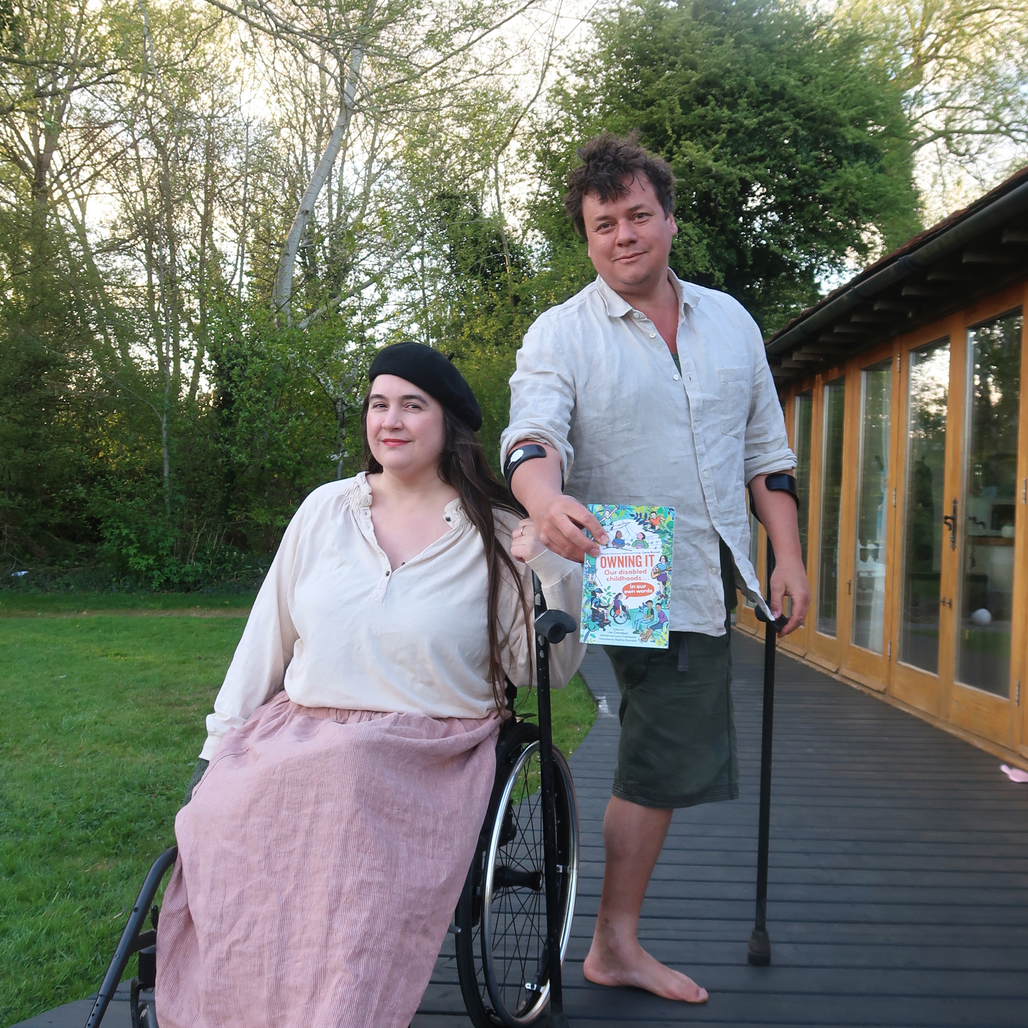 Lucy and James Catchpole - a white, disabled couple, holding 'Owning It. Lucy is in her wheelchair, James has one leg and crutches.