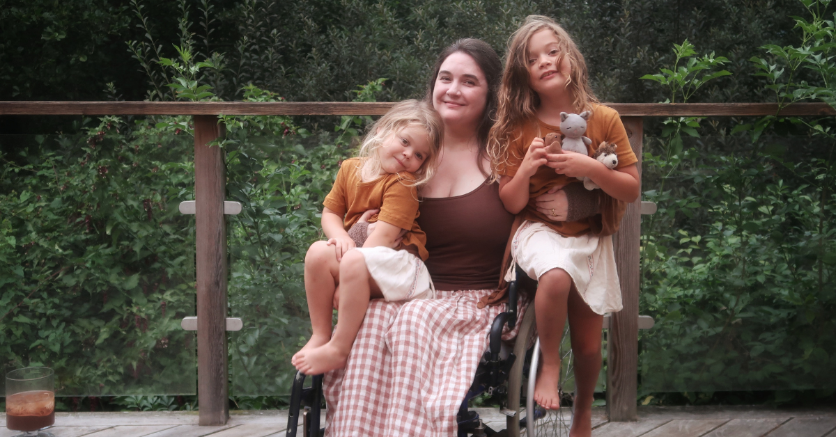 Lucy - a white woman with long brown hair - sits in her wheelchair, with both daughters perched on and around her. They're smiling at the camera. 