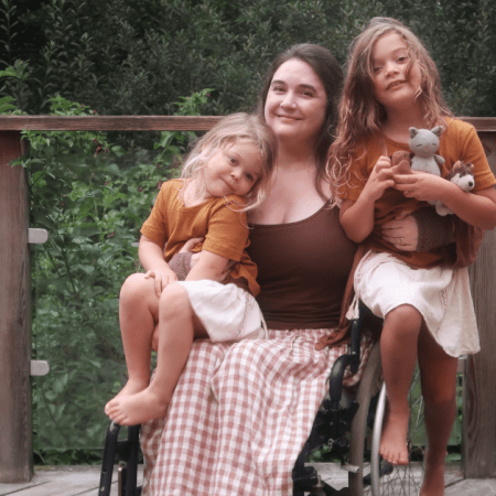 Lucy - a white woman with long brown hair - sits in her wheelchair, with both daughters perched on and around her. They're smiling at the camera.